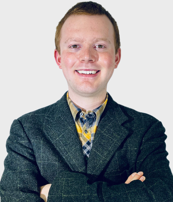 An image of Josh, a man with short ginger hair, who is smiling at the camera, he is wearing a green tweed jacket and has his arms folded